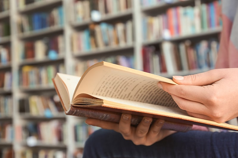 A close up of an open book in someones hand while they are reading in a library