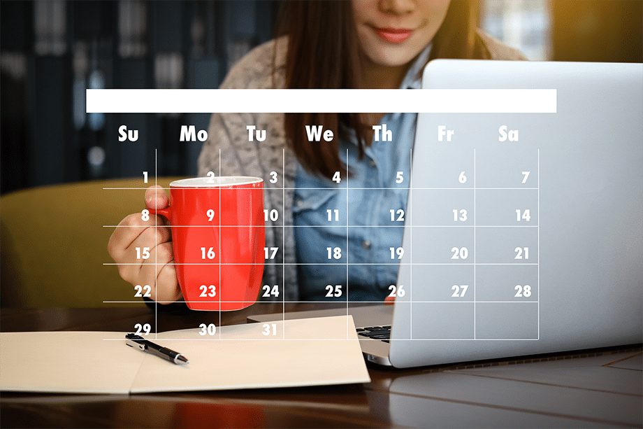 A woman sitting at a wooden desk in front of her laptop while holding a red coffee mug. The image has a graphic of a blank calendar over the photo of the woman working