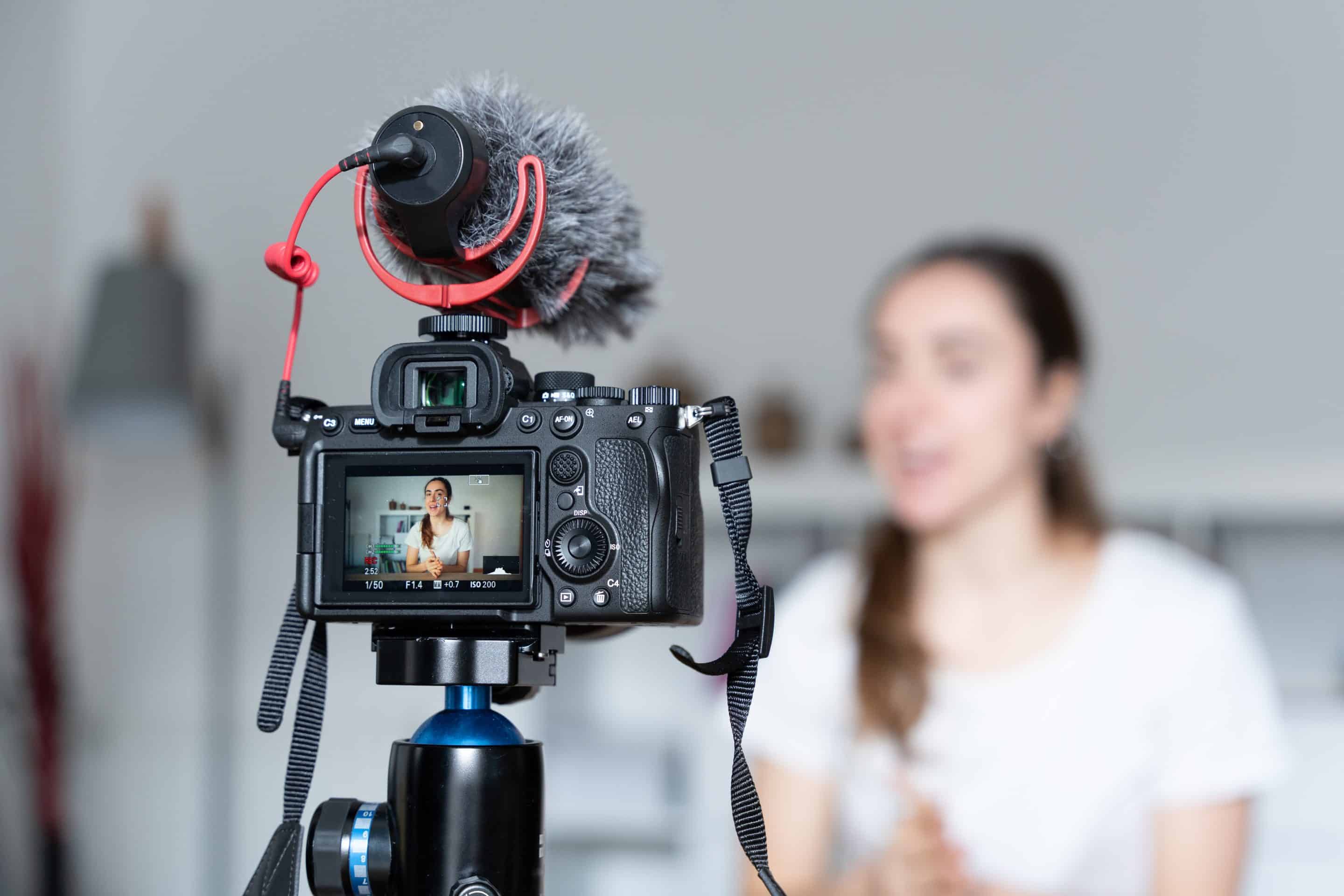 A photo of a woman sitting at a desk in front of a video camera with a mic attached to the top. The video camera is the only element of the photo in focus, the rest is blurred