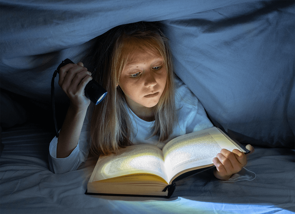 A young girl reading a book under her bed covers using a flashlight to read